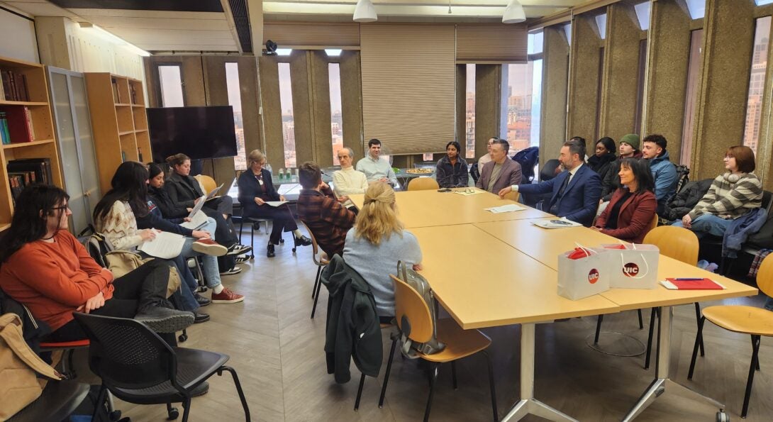 Approximately twenty people sit around a rectangular table, listening to a speaker who is reading from a paper.